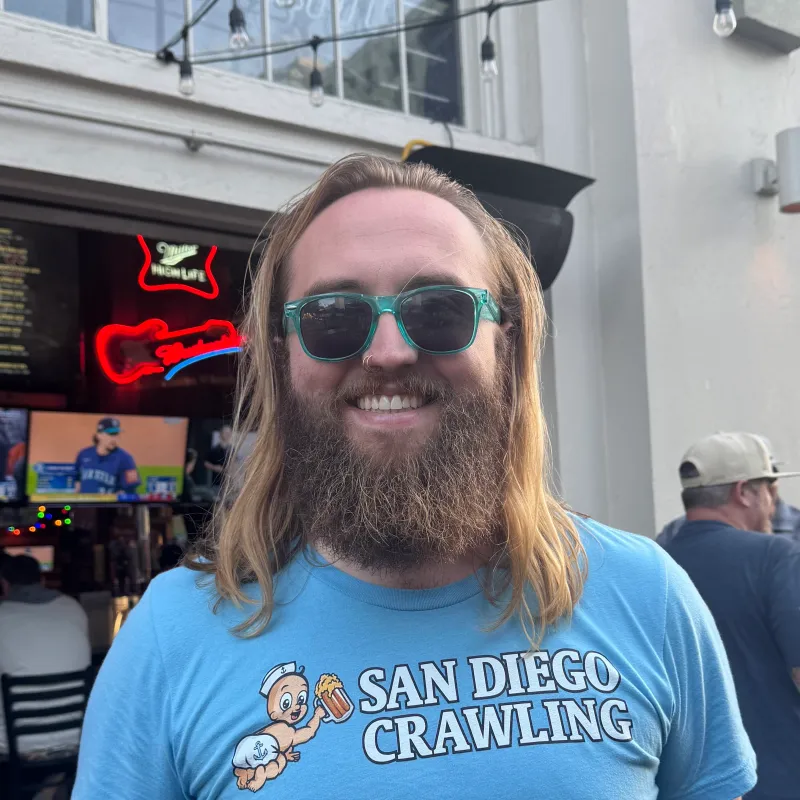 17610542628135-IMG_2558 Man with long hair and beard wearing sunglasses and a 'San Diego Crawling' shirt in front of a bar.